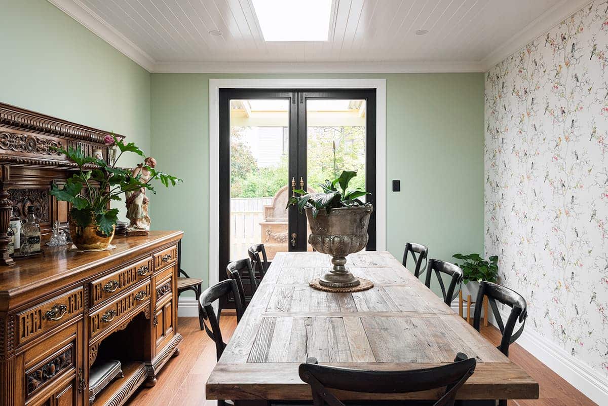 A dining room with a wooden table, six chairs, an ornate sideboard, potted plants, floral wallpaper, and glass doors leading outside.