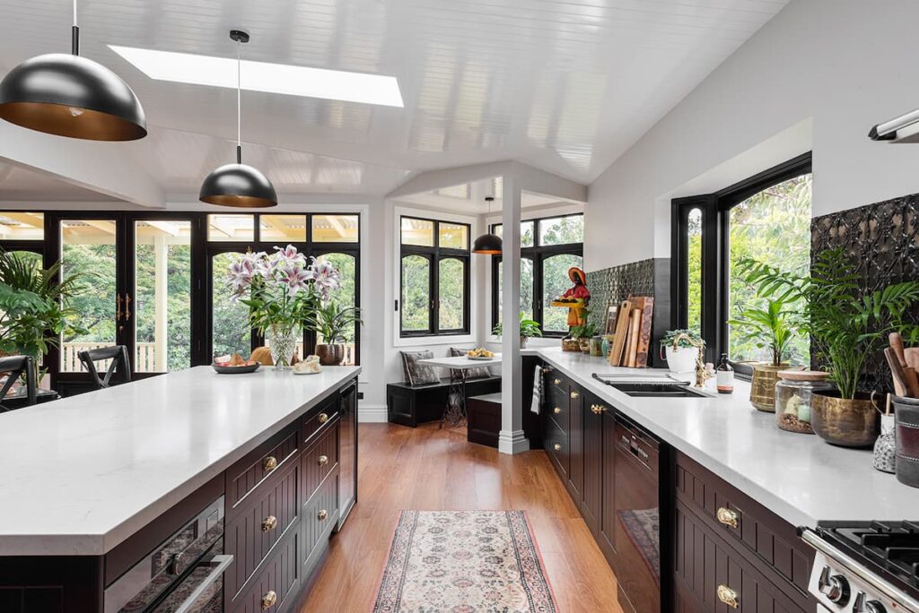 Modern kitchen with dark cabinets, white countertops, wood flooring, pendant lights, large windows, plants, and a small seating area by the window.