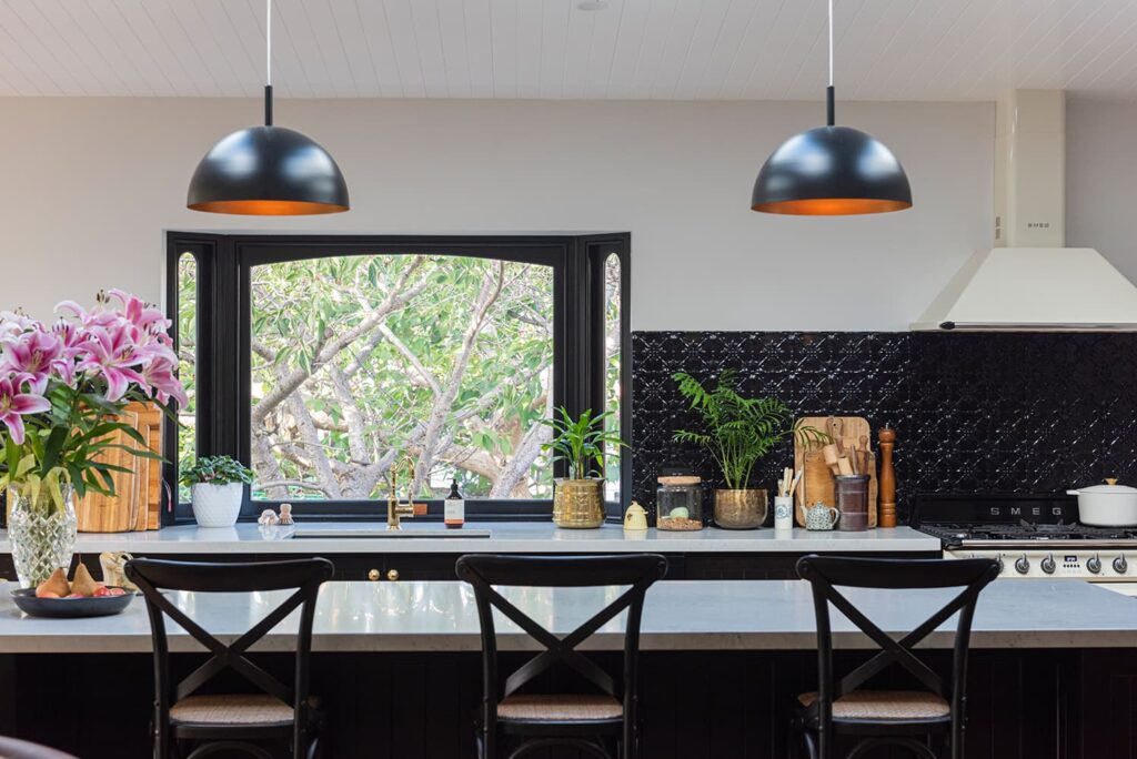 Modern kitchen with a marble island, black chairs, two pendant lights, a large window, black backsplash, and various plants and decor items on the counter.