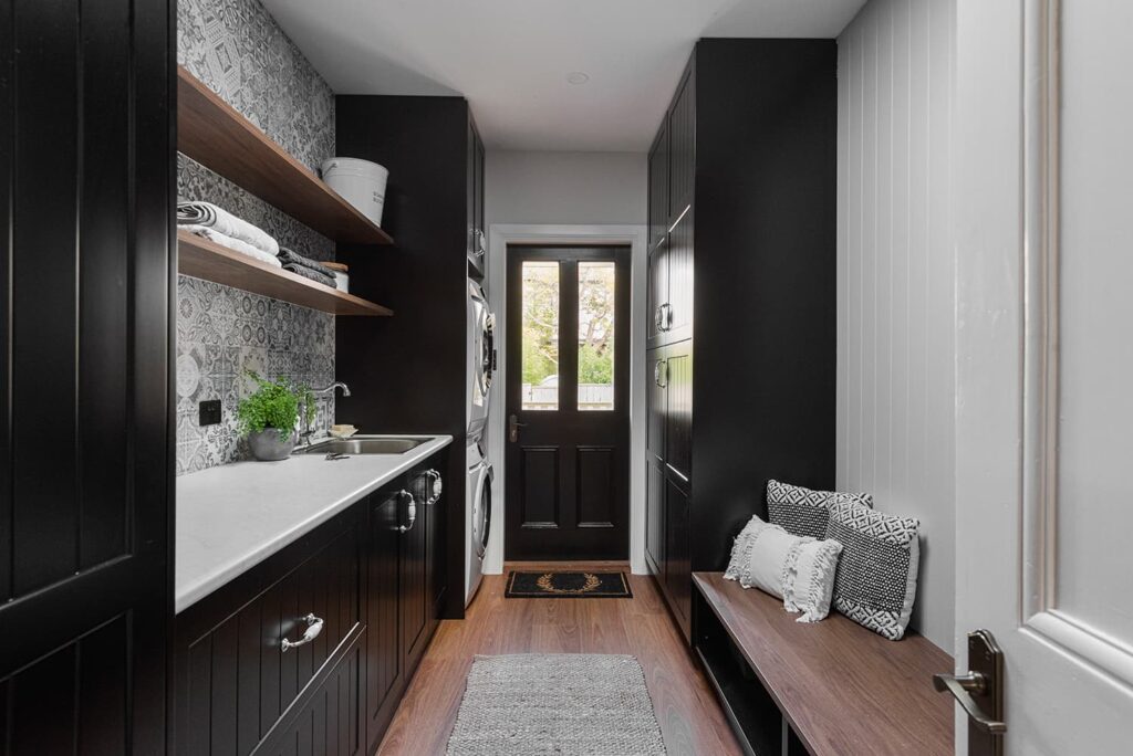 A modern laundry room with black cabinets, patterned backsplash, open shelves, stacked washer and dryer, and a bench with cushions near a glass-panel door leading outside.