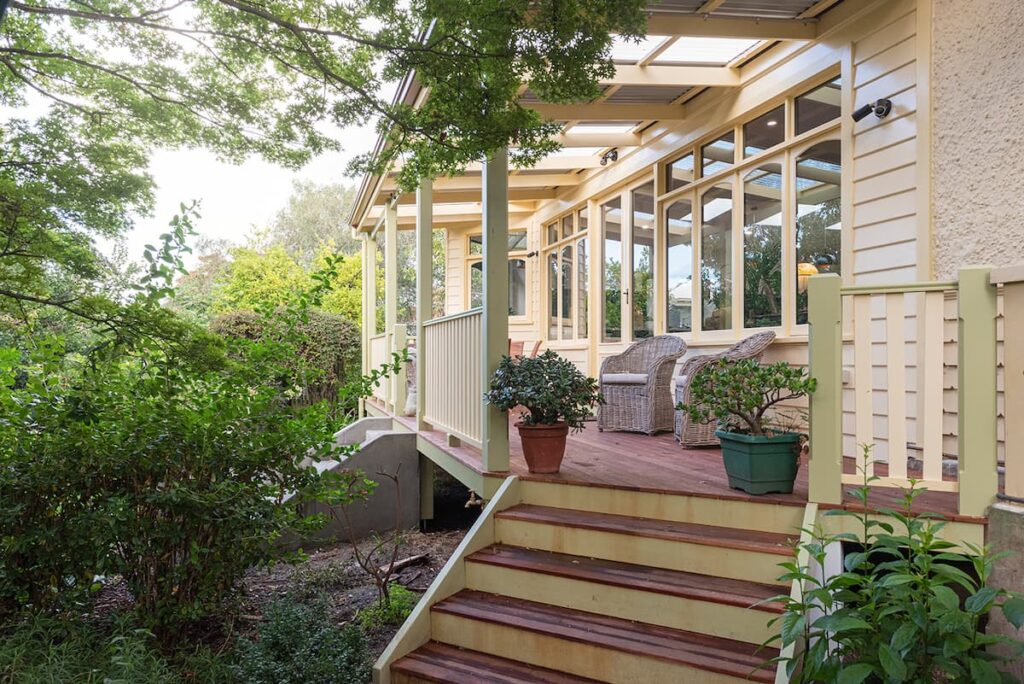 Wooden porch with steps, potted plants, and wicker chairs, attached to a light-colored house with large windows, surrounded by greenery.
