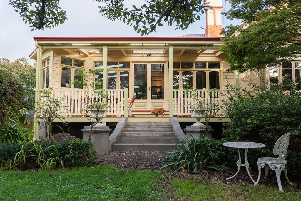 A cream-colored house with large windows and a covered porch, stone steps leading to a garden with greenery, and a white metal table and chairs in the foreground.