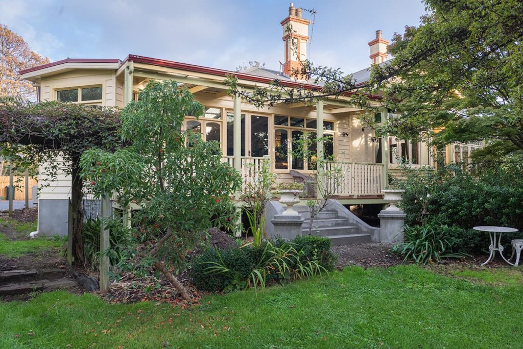 Large, light-colored house with a front porch, surrounded by greenery and trees, with concrete steps leading up to the entrance and an outdoor table and chairs on the lawn.