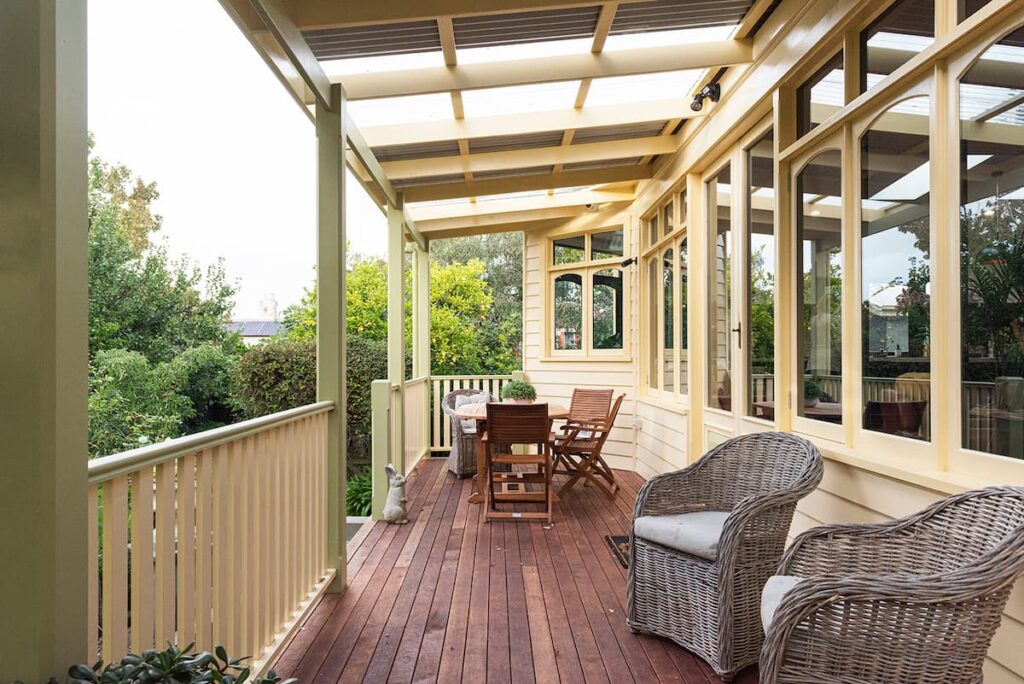 A covered wooden porch with wicker chairs, a wooden dining table set, and railings, overlooking a green garden area.