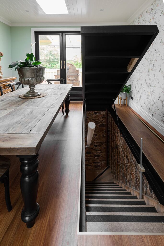 A wooden dining table sits beside an open staircase leading down to a stone-walled basement, with double doors and potted plants in the background.