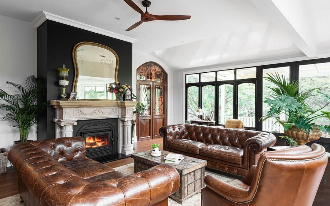 A living room with brown leather sofas, a matching armchair, a wooden coffee table, fireplace with a mirror above, houseplants, and large windows overlooking greenery.