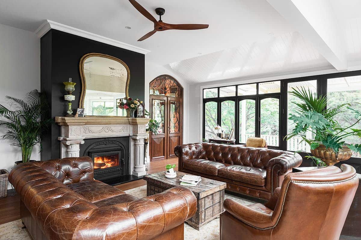 A living room with brown leather sofas, a matching armchair, a wooden coffee table, fireplace with a mirror above, houseplants, and large windows overlooking greenery.