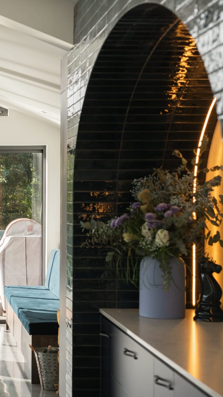 Modern living space with a suspended black fireplace, large windows, outdoor view, built-in bench seating, and a kitchen area with a flower arrangement on the counter.