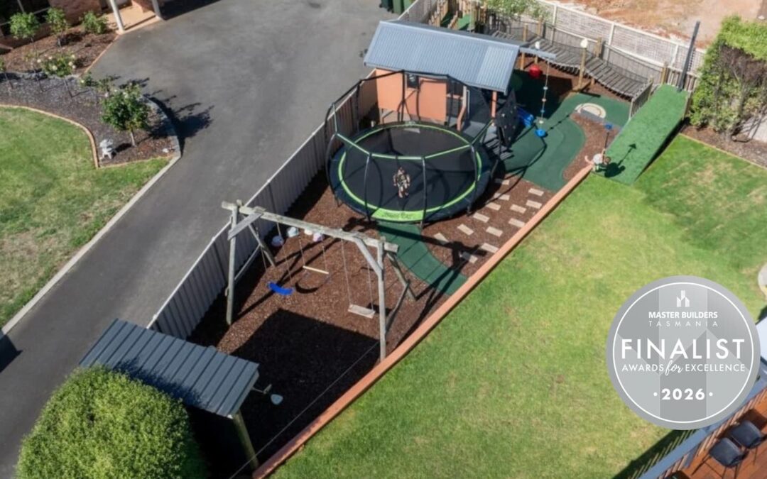 Aerial view of a backyard with swing set, trampoline, covered play area, stepping stones, garden beds, and a circular emblem on the lawn that reads “Finalist 2026.”.